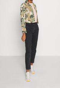 Camo-mönstrad kort jacka i grön, beige och brun; ljusrosa topp; högmidjade svarta jeans; grå och gula sneakers.