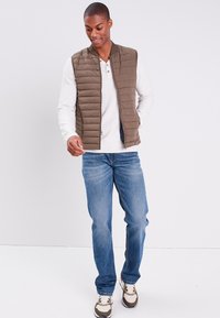 Braun gesteppte Weste über einem hellen Langarmhemd, kombiniert mit blauen Jeans und mehrfarbigen Sneakers; entspannte Passform, Reißverschluss, und Taschen.