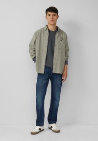 Man in een grijze T-shirt, beige geruite overhemd met knopen, blauwe jeans en witte sneakers met zwarte strepen, staande tegen een effen achtergrond.