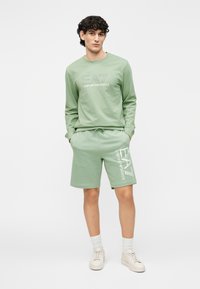Conjunto de sudadera verde y pantalones cortos en una mezcla suave de algodón. Presenta el logo EA7 en ambas piezas, con un ajuste relajado y cintura con cordón.