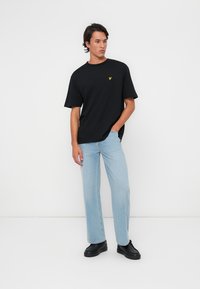 Camiseta negra de manga corta con un pequeño logotipo amarillo, combinada con jeans de denim azul claro y calzado negro. Diseño minimalista, corte relajado.
