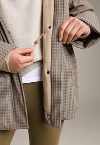 Handen die een beige en bruin geruit jack met elastisch trekkoord aanpassen, gedragen over een licht beige trui en olijfgroene broek.