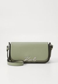 Zelená kožená kabelka přes rameno s klopou, stříbrným logem a nastavitelným popruhem. Hladká textura, strukturovaný tvar.