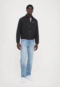 Black zip-up jacket with a collar, paired with light blue jeans and white sneakers. The jacket features a subtle logo on the chest.