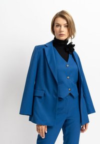Blauer maßgeschneiderter Anzug mit einem Blazer mit breiten Revers, einer passenden Weste mit drei Knöpfen und maßgeschneiderten Hosen. Darunter ein schwarzer Rollkragenpullover.