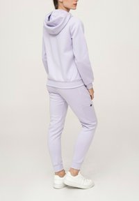 Ensemble de survêtement lavande clair comprenant une veste à capuche et un pantalon ajusté avec des poches latérales. Le matériau est doux avec une texture lisse, associé à des baskets blanches.
