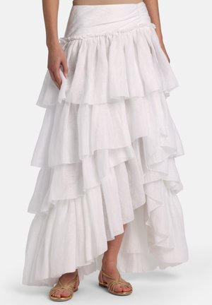 Femme portant une jupe maxi blanche à volants superposés associée à des sandales plates dorées à lanières, debout devant un fond uni.