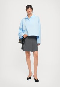 Vrouw in een lichtblauwe oversized blouse, een grijze geplooide rok, zwarte hakken en met een zwart, gewatteerd schoudertasje met een gouden kettingband.