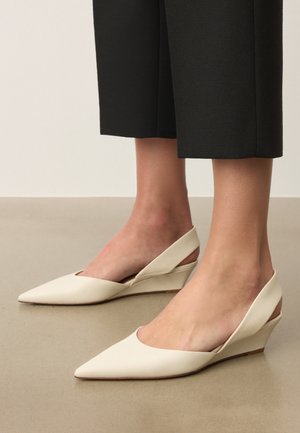 Jalad, mis kannavad kreemikas värvi terava ninaga sling-back kiilkontsaga jalanõusid, kokku sobitatud mustade lühendatud pükstega, tasasel põrandal.