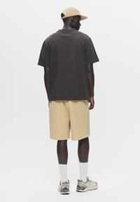 Black short-sleeve shirt, beige shorts, and gray sneakers with white accents. A tan cap completes the outfit, viewed from the back.