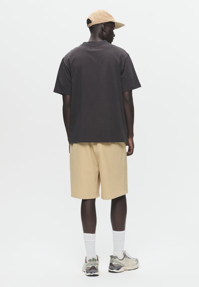 Zwarte korte mouwen shirt, beige shorts en grijze sneakers met witte accenten. Een tan pet maakt de outfit compleet, gezien vanaf de achterkant.