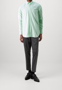Camisa de botones verde claro con cuello, combinada con pantalones de vestir grises y zapatos negros. La camisa presenta un pequeño logo en el pecho.