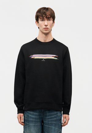 Νεαρός με καστανά μαλλιά, φορώντας μαύρο φούτερ με πολύχρωμο οριζόντιο σχεδιασμό από πινελιές και μπλε τζιν, στέκεται μπροστά σε ένα απλό φόντο.