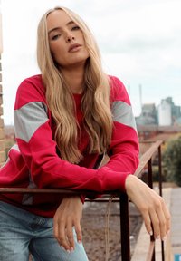 Ragazza giovane con lunghi capelli biondi che indossa una felpa rossa e grigia, si appoggia a una ringhiera metallica arrugginita all'aperto, con uno sfondo urbano.