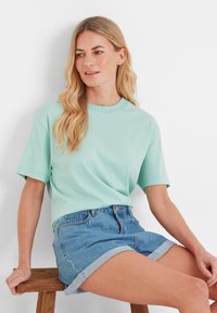 Woman with long blonde hair wearing a mint green t-shirt and rolled-up denim shorts sitting on a wooden stool against a white wall.