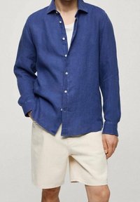 Chemise en lin bleue à boutons, manches longues, texture de tissu légère et boutons blancs, associée à un short de couleur crème.