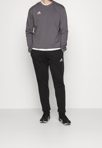 Sweat-shirt gris à manches longues avec un logo Adidas, assorti à un pantalon de survêtement noir arborant le logo Adidas. La personne porte des chaussures de sport noires.