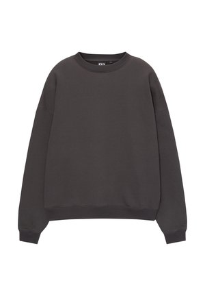 Personne portant un sweat-shirt noir ample, un jean délavé clair et deux colliers de perles superposés, debout avec une main dans la poche.