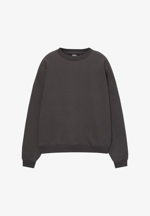 Person iført en løs sort sweatshirt, lyseblå jeans og to lagdelte perlehalskæder, stående med den ene hånd i lommen.