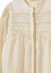 Blusa color crema realizzata in tessuto leggero, con chiusura a bottoni, strisce di pizzo in rilievo e texture arricciata in vita.