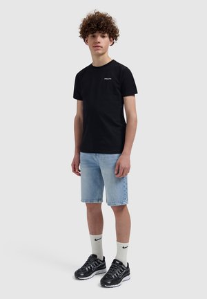 Tienerjongen met krullend haar, gekleed in een zwart T-shirt, lichtblauwe denimshorts, witte Nike-sokken en zwarte Nike-sneakers, staand tegen een effen achtergrond.