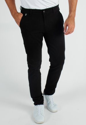 Pantalon en coton noir avec une coupe slim, comportant une fermeture à bouton à l'avant, des passants de ceinture et des poches latérales. Associé à des baskets blanches.