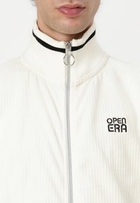 Weißes, geripptes Zip-Jacket mit hohem Kragen, schwarzen Streifen und silbernem Reißverschluss, auf der Brust mit "OPEN ERA" bestickt.