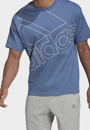 Man wearing blue Adidas t-shirt with large white outlined logo and light gray Adidas sweatpants standing with arms slightly bent.