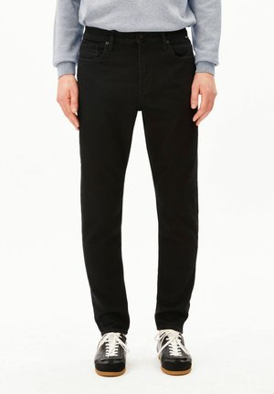 Persoon draagt strakke zwarte broek, grijze sweatshirt en zwart-witte veter sneakers, staand tegen een witte achtergrond.