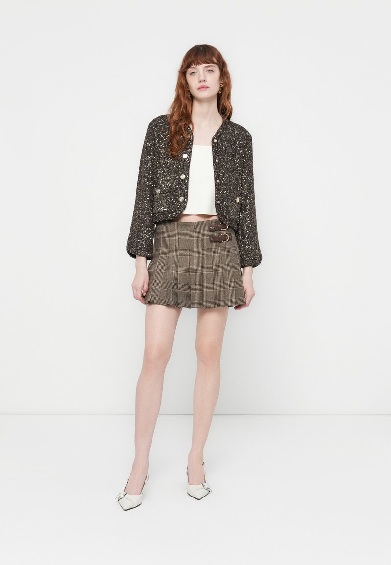 Junge Frau trägt eine schwarze, mit Pailletten besetzte, cropped Jacke, ein weißes Crop-Top, einen braunen, karierten, gefalteten Minirock mit Schnallen und weiße High Heels.