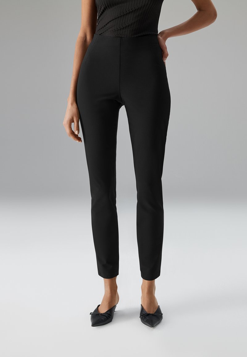 Zwarte nauwsluitende broek met een gladde textuur, hoge taille, taps toelopend ontwerp en zijzakken, gecombineerd met zwarte puntige platte schoenen.
