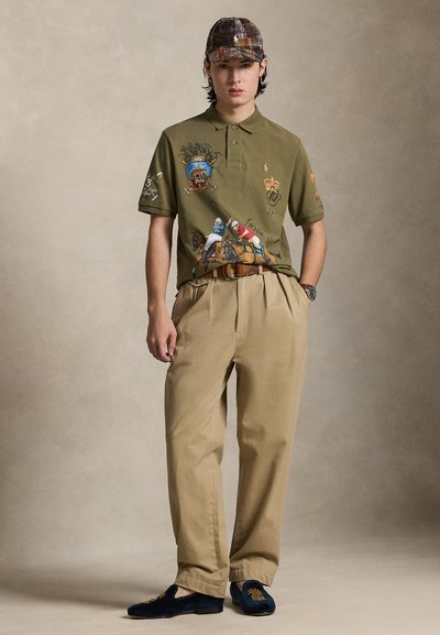 Πόλο πουκάμισο ελιάς με κεντημένα σχέδια, μπεζ πλισέ παντελόνια και ναυτικό σκούρα loafers με χρυσό έμβλημα. Καπέλο μπέιζμπολ με πολύχρωμο μοτίβο.