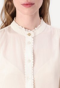 Light beige blouse with a ruffled collar, featuring a central button placket, transparent buttons, and scalloped edges. Soft fabric texture.