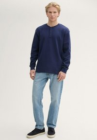Marineblaue Langarm-Henley-Shirt mit drei Knöpfen, kombiniert mit hellblauen klassischen Jeans und dunklen Sneakern, das ein einfaches Design aufweist.