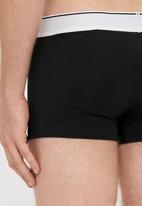 Melnas kokvilnas bokseršorti ar baltu elastīgu jostu, uz kuras ir plāna melna svītra. Audums izskatās gluds un pieguļošs.