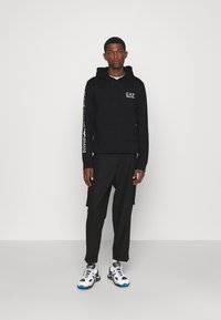 Felpa nera con tasca frontale e logo, abbinata a pantaloni cargo neri. Il modello indossa scarpe sportive bianche e blu. Design minimalista, texture liscia.