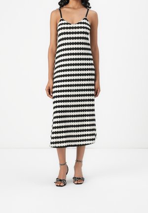 Femme portant une robe midi sans manches à rayures horizontales noires et blanches avec de fines bretelles, et des sandales noires avec des brides de cheville cloutées.