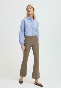 Camisa de botones a rayas azules con un frente fruncido, combinada con pantalones acampanados marrones y zapatos planos marrones, sobre un fondo claro.