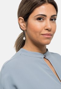 Silberne Tropfohrringe mit einer strukturierten Kette und einem wei�ßen Perlenakzent. Die Bluse ist hellblau mit einem Stehkragen und einem Schlüsselanschnitt.