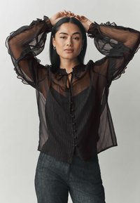 Blusa nera in chiffon con colletto e polsini arricciati, caratterizzata da righe verticali e chiusura con bottoni. Abbinata a pantaloni scuri texturizzati.