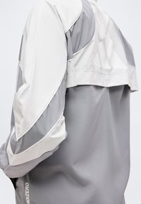 Gros plan d'une veste de sport grise et blanche avec le logo "ADIZERO" sur la manche, présentant un tissu superposé et des détails de coutures courbes.