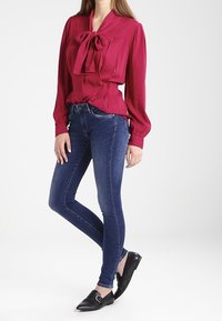 Långärmad bordeauxblus med en slipsdetalj, ihopparad med mörkblå slim fit-jeans och svarta platta skor. Slät tygtextur.