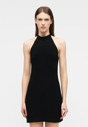 Femme aux cheveux bruns raides portant une robe noire sans manches à col licou, debout devant un fond clair uni.