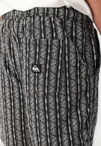 Shorts à motifs noir et blanc présentant des rayures verticales, des motifs floraux, une taille élastique et des poches latérales. Comprend un patch logo.