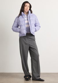 Chaqueta acolchada lavanda con patrón de acolchado horizontal, combinada con un suéter a rayas en tonos apagados y pantalones grises con rayas. Calzado negro.