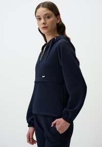 Felpa con cappuccio blu navy, dotata di cordino e zip dorata. Presenta maniche lunghe, leggermente affusolate con polsini elasticizzati e una tasca frontale.