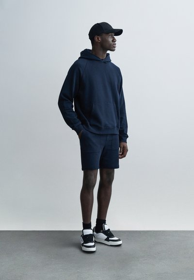 Tmavě modrá mikina s kapucí a klokaní kapsou, doplněná tmavě modrými kraťasy. Bílé a černé tenisky, černá čepice s logem. Hladká textura, ležérní design.