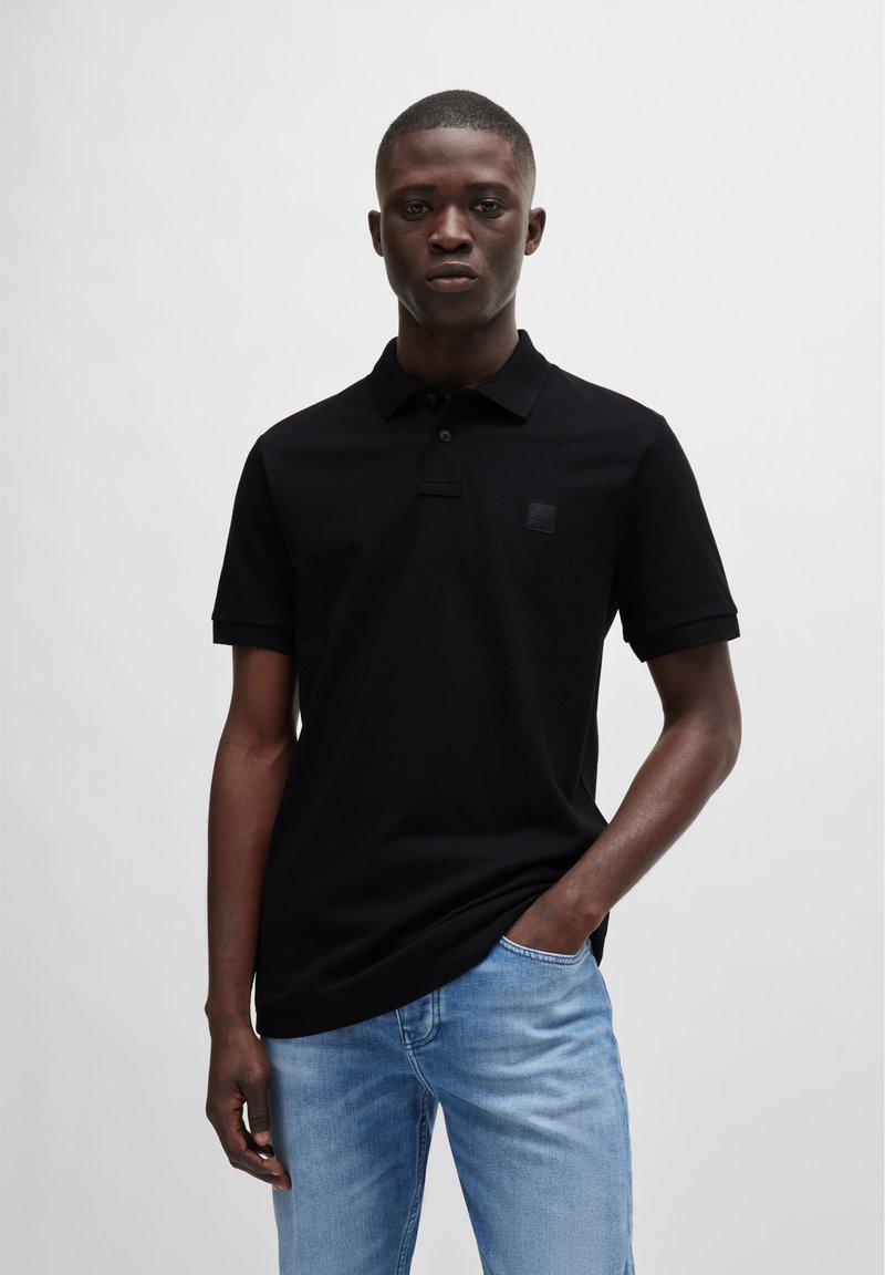 Schwarzes Poloshirt mit kurzen Ärmeln, das mit einem dezenten Logo-Patch auf der Brust verziert ist, kombiniert mit hellblauen Jeans aus Denim. Glatte Textur.