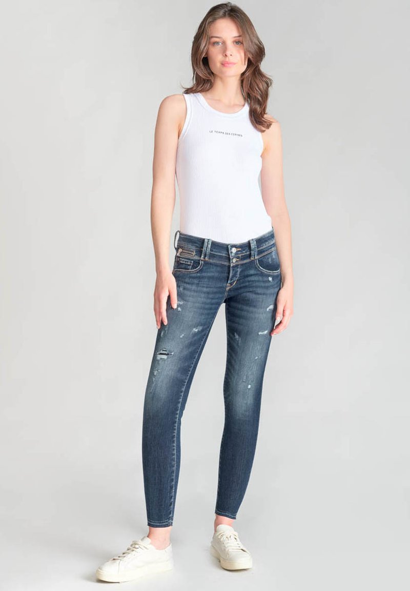 Jeans ajustados en denim azul oscuro con detalles desgastados y una cintura de altura media, combinados con una blusa sin mangas blanca y zapatillas blancas.