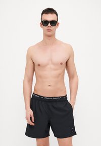 Calções de banho pretos com uma cintura elástica, apresentando texto branco. Modelo a usar óculos de sol escuros, fundo é branco liso.